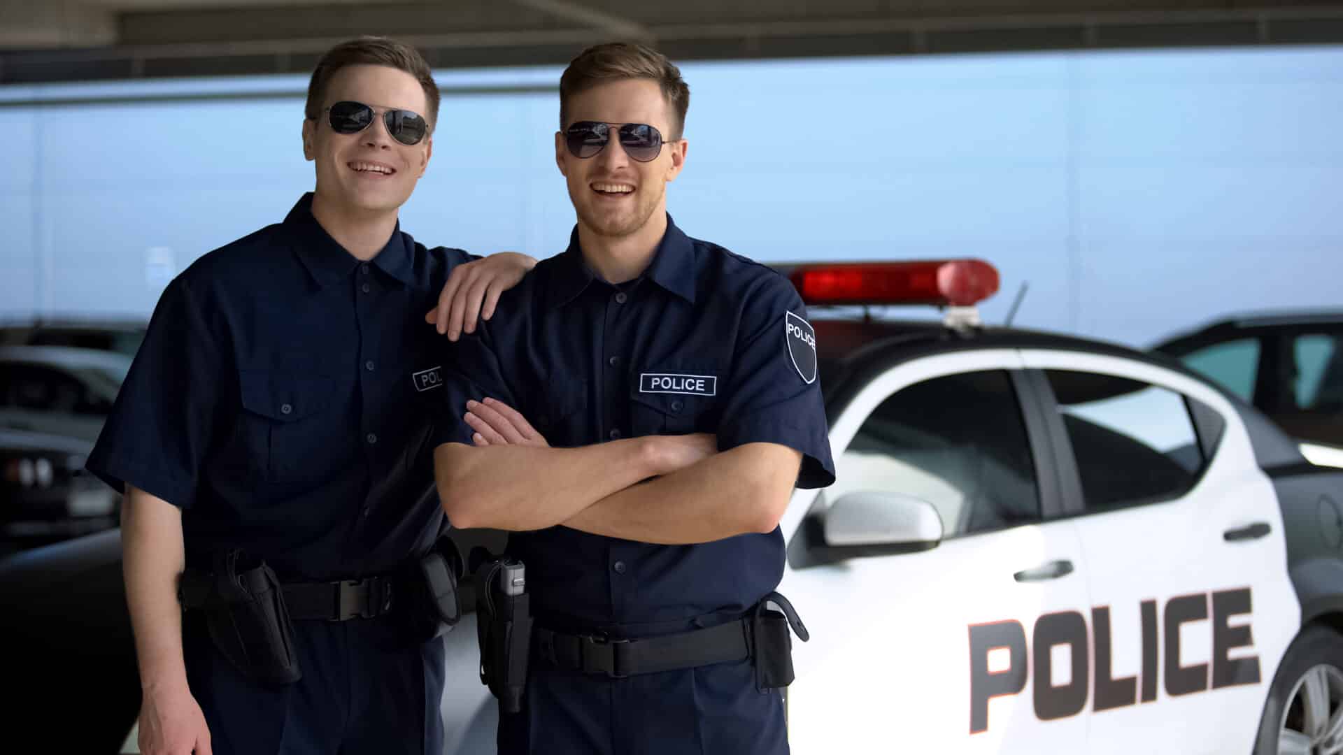 Cheerful police mates in sunglasses smiling into camera against squad car, work